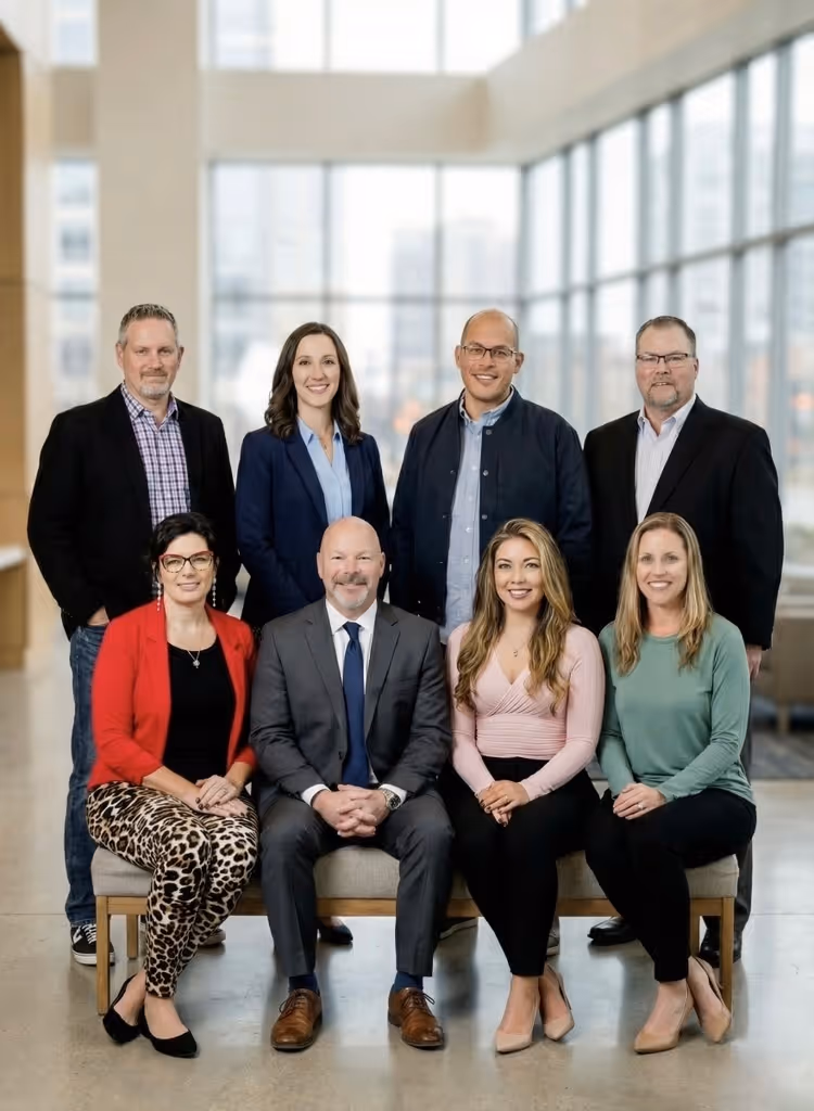 Group portrait of eight professionally dressed people posing in an office with large windows.