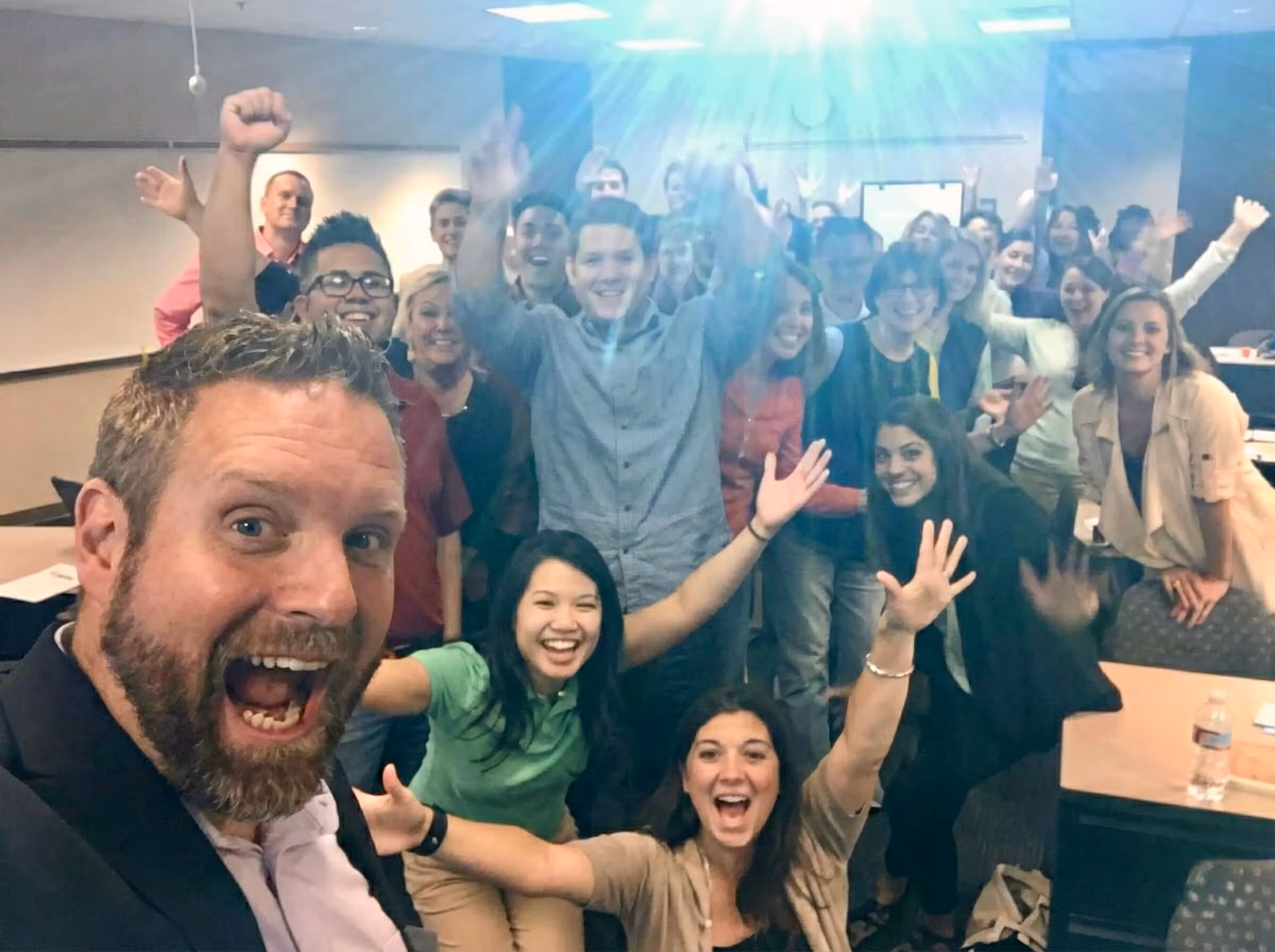 Large group of smiling people in a classroom, some raising hands, posing for a lively selfie.