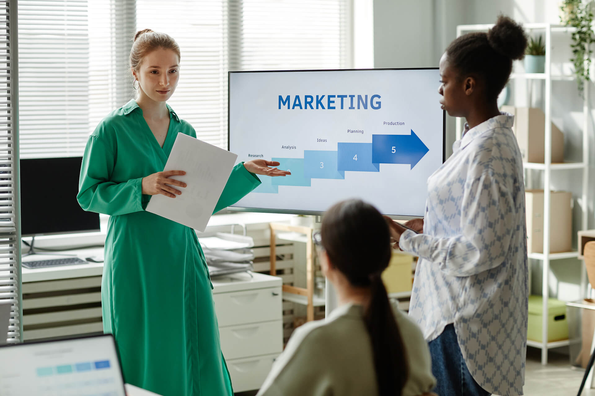 Two women present a marketing plan using a screen showing stages from research to production.