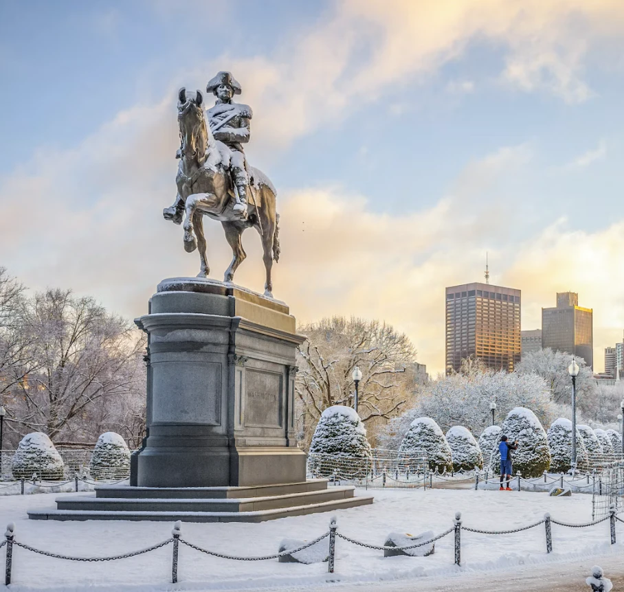 Snow-covered equestrian statue in a city park with leafless trees and buildings in the background.